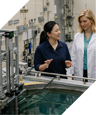 Two women, one in a lab coat, talk while looking at an experiment monitoring a nuclear experiment