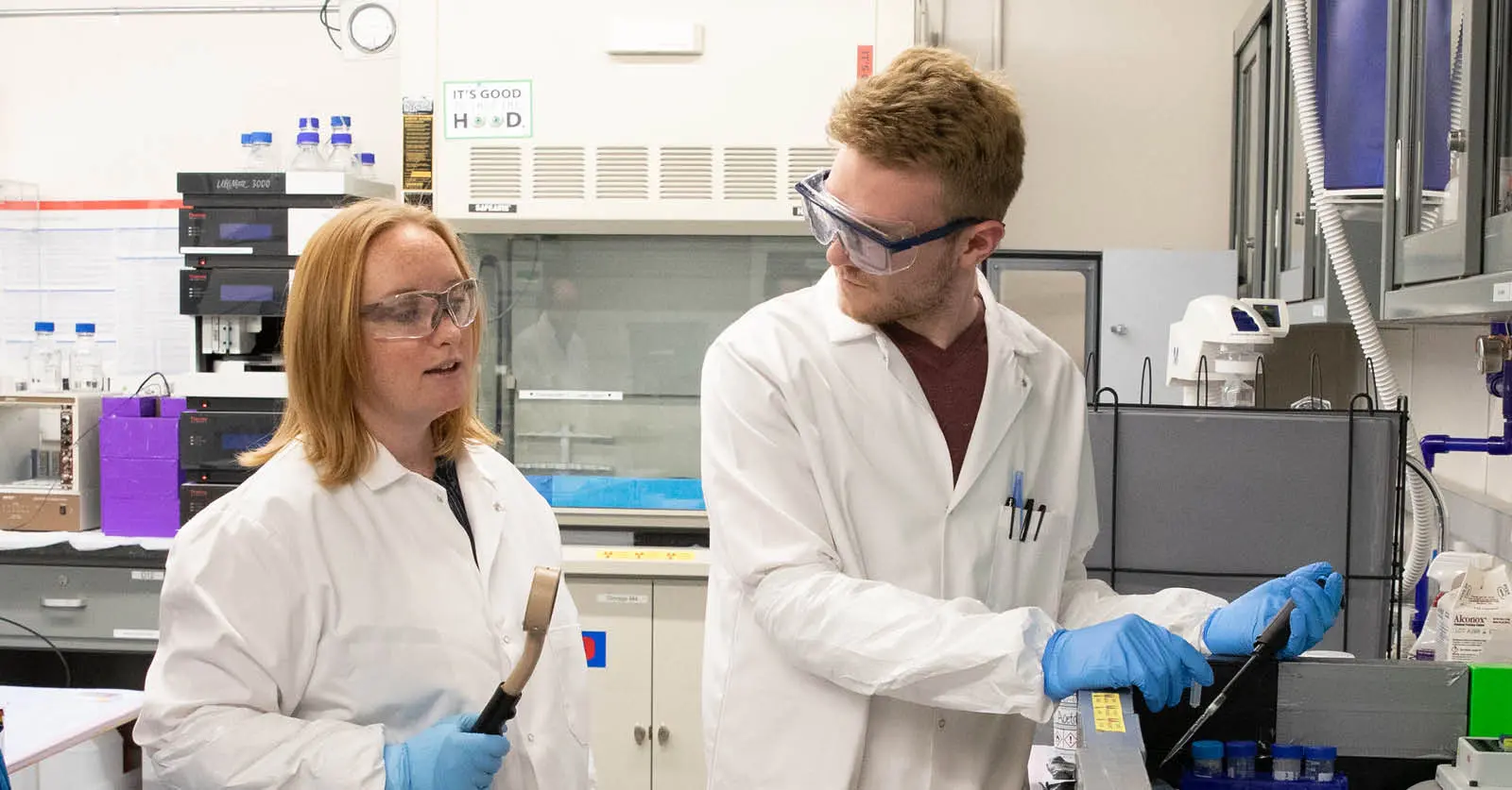 Two college-age students in lab coats, taking samples and speaking together